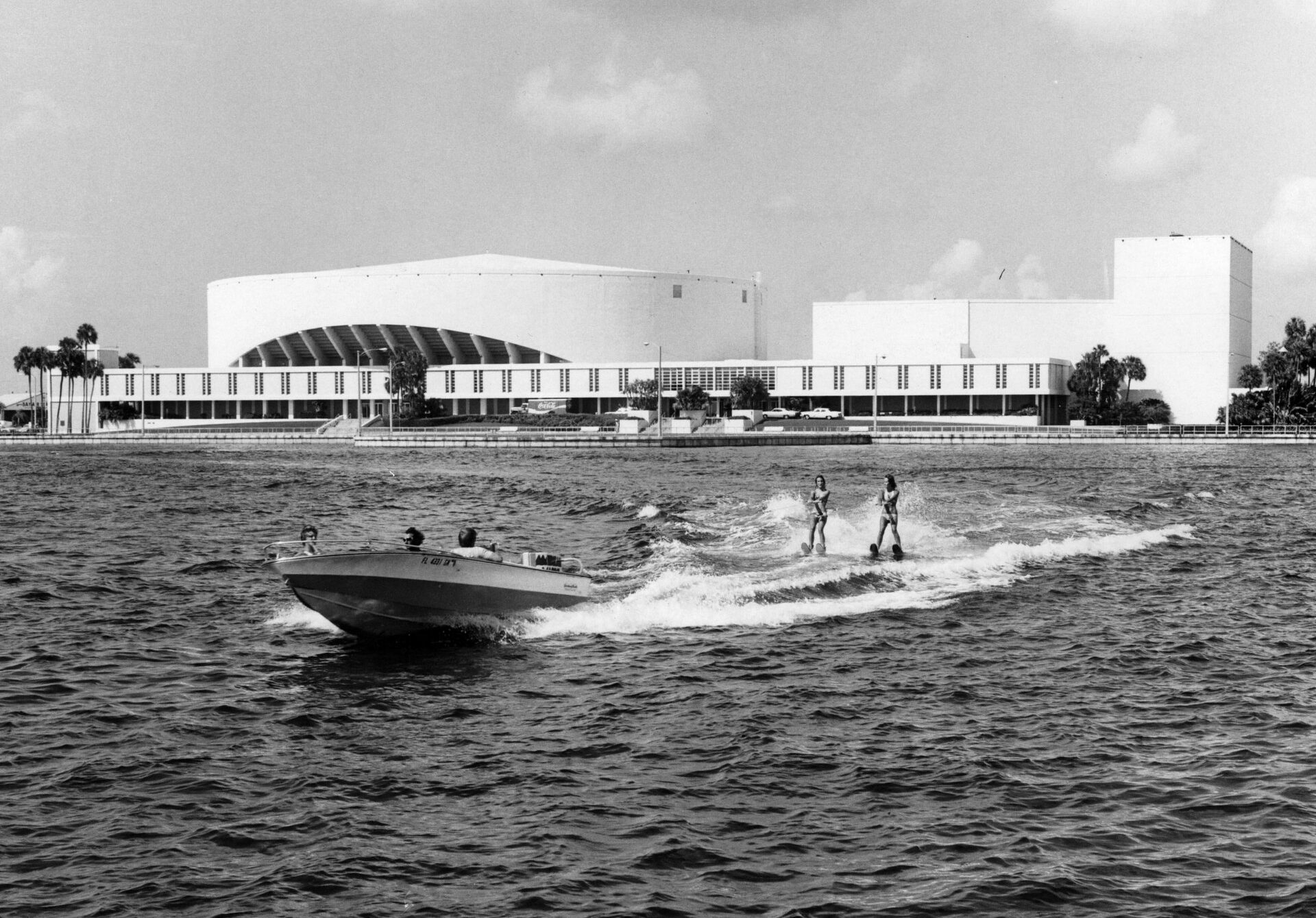 Two water skiers towed by a motorboat on a bay, with a large modernist arena building visible on the shoreline behind them