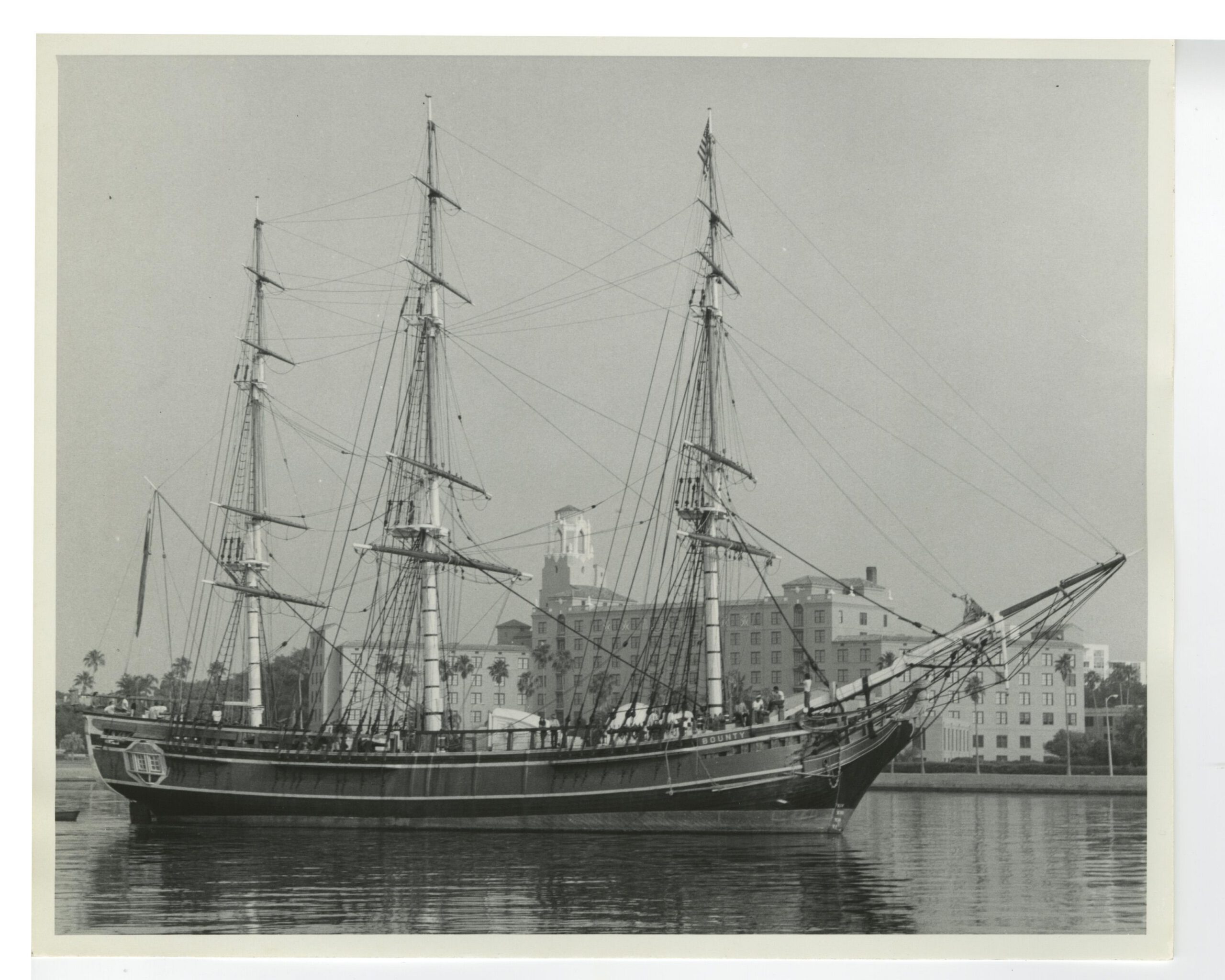 Black-and-white photo of the tall ship Bounty moored in a harbor with large buildings visible in the background