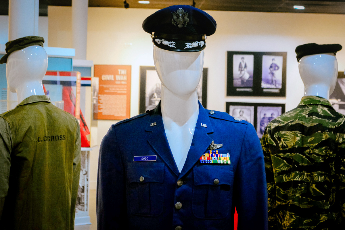 Museum display of military uniforms on mannequins, including a U.S. Air Force dress uniform with ribbons and pilot wings, an olive drab jacket labeled C. Cross, and a camouflage shirt