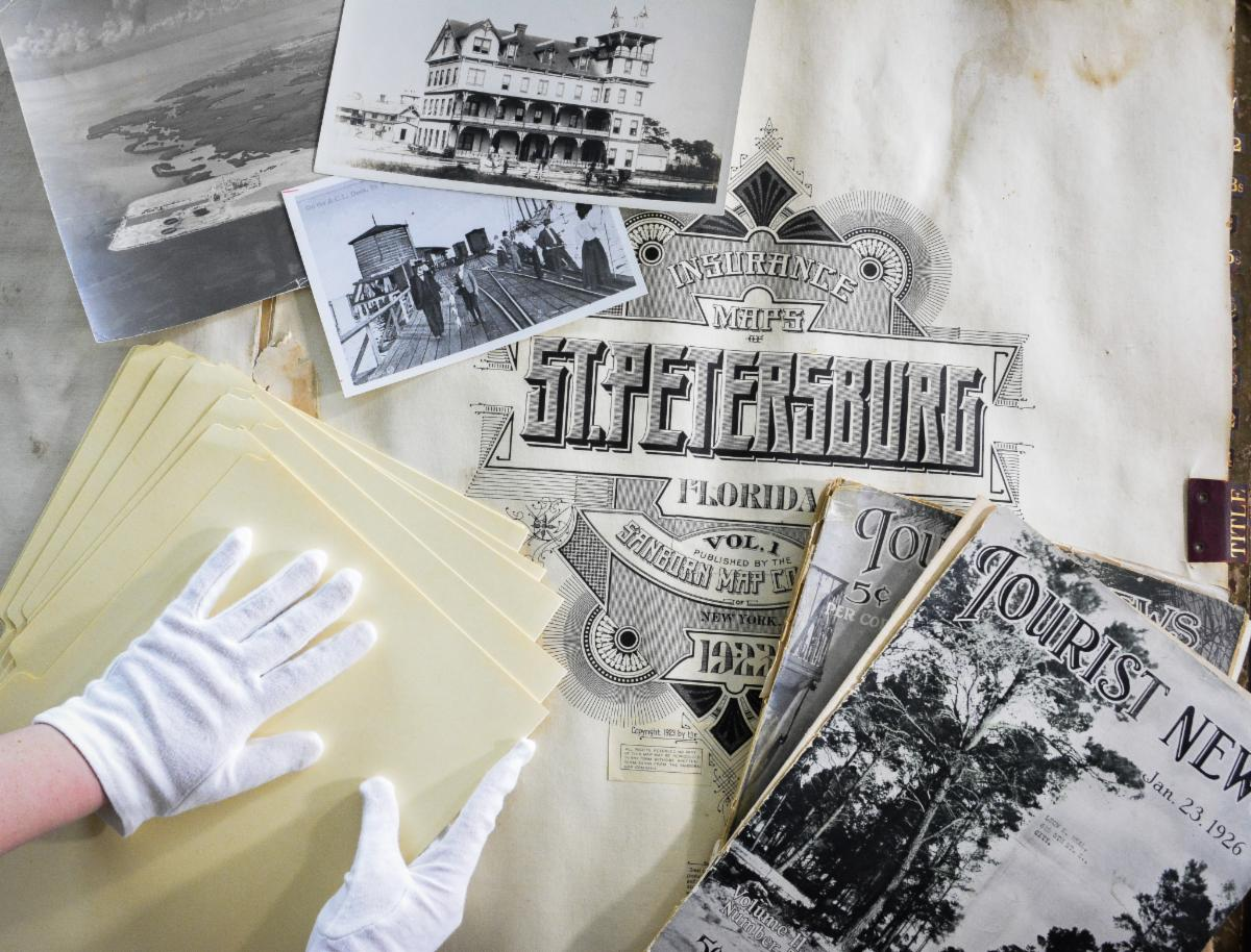 Gloved hands handling historical St. Petersburg, Florida archival materials including Sanborn insurance maps, vintage photographs, and Tourist News publications from the 1920s