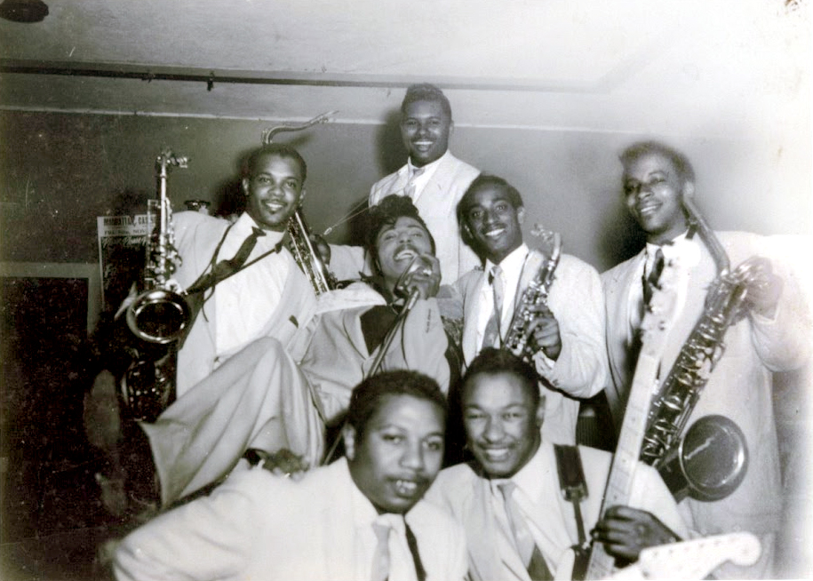 Vintage black-and-white photo of a jazz or R&B band posing together, several members holding saxophones and a guitar, all dressed in light suits