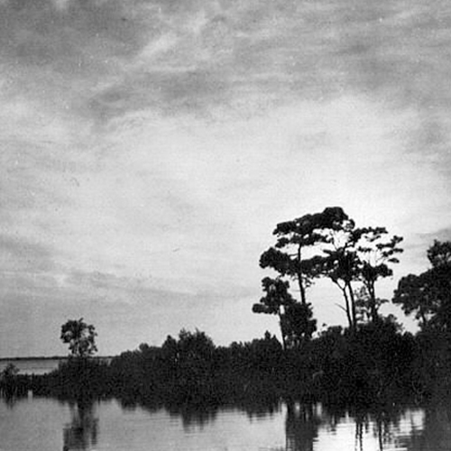 Black-and-white photograph of a calm waterway lined with trees reflected in the water under a cloudy sky