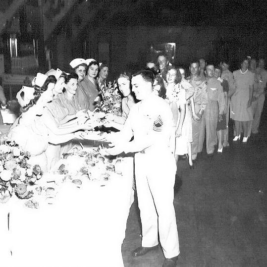 A soldier in uniform receives items from nurses at a table during a World War II-era event, with a line of people behind him