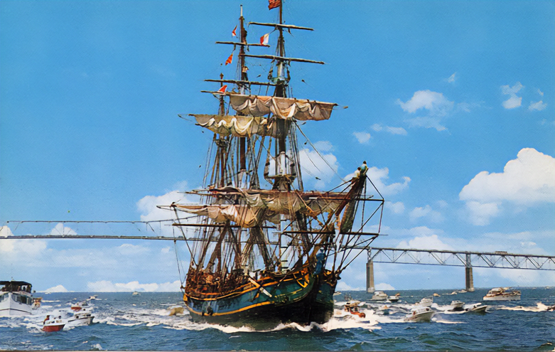 Tall ship with furled sails sailing through open water, surrounded by small motorboats, with a bridge visible in the background