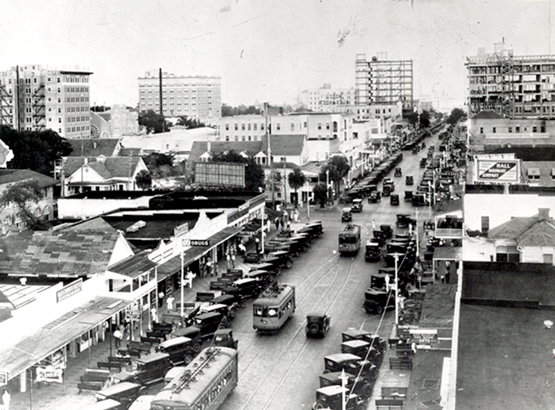 Aerial black-and-white photo of a busy early 20th-century city street with streetcars, early automobiles, and storefronts lining both sides