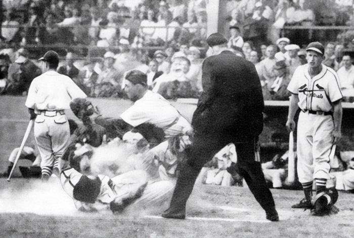Vintage black-and-white photo of a baseball player sliding into home plate in a cloud of dust as the catcher attempts a tag, with an umpire and Cardinals players nearby