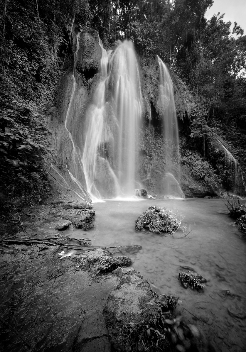 Black and white long-exposure photograph of a tall waterfall cascading over mossy rocks into a shallow pool surrounded by forest