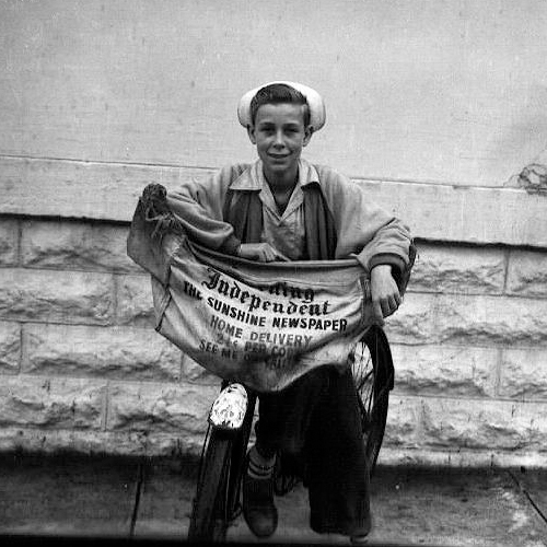 Young newspaper delivery boy on a bicycle holding a canvas bag reading Independent The Sunshine Newspaper Home Delivery