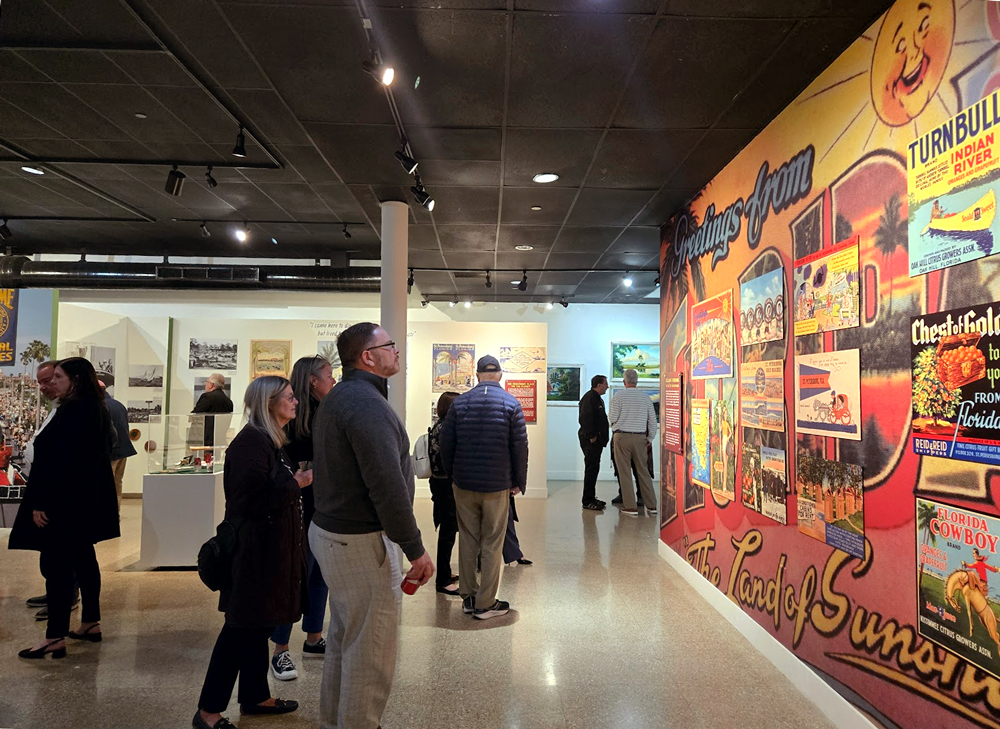 Visitors exploring a museum exhibit featuring a large colorful mural reading Greetings from the Land of Sunshine with vintage Florida imagery and citrus brand labels