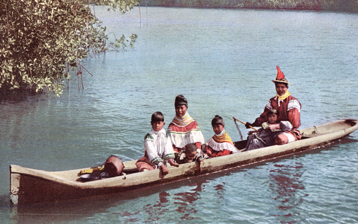 Seminole family in traditional dress seated in a dugout canoe on calm water near mangroves