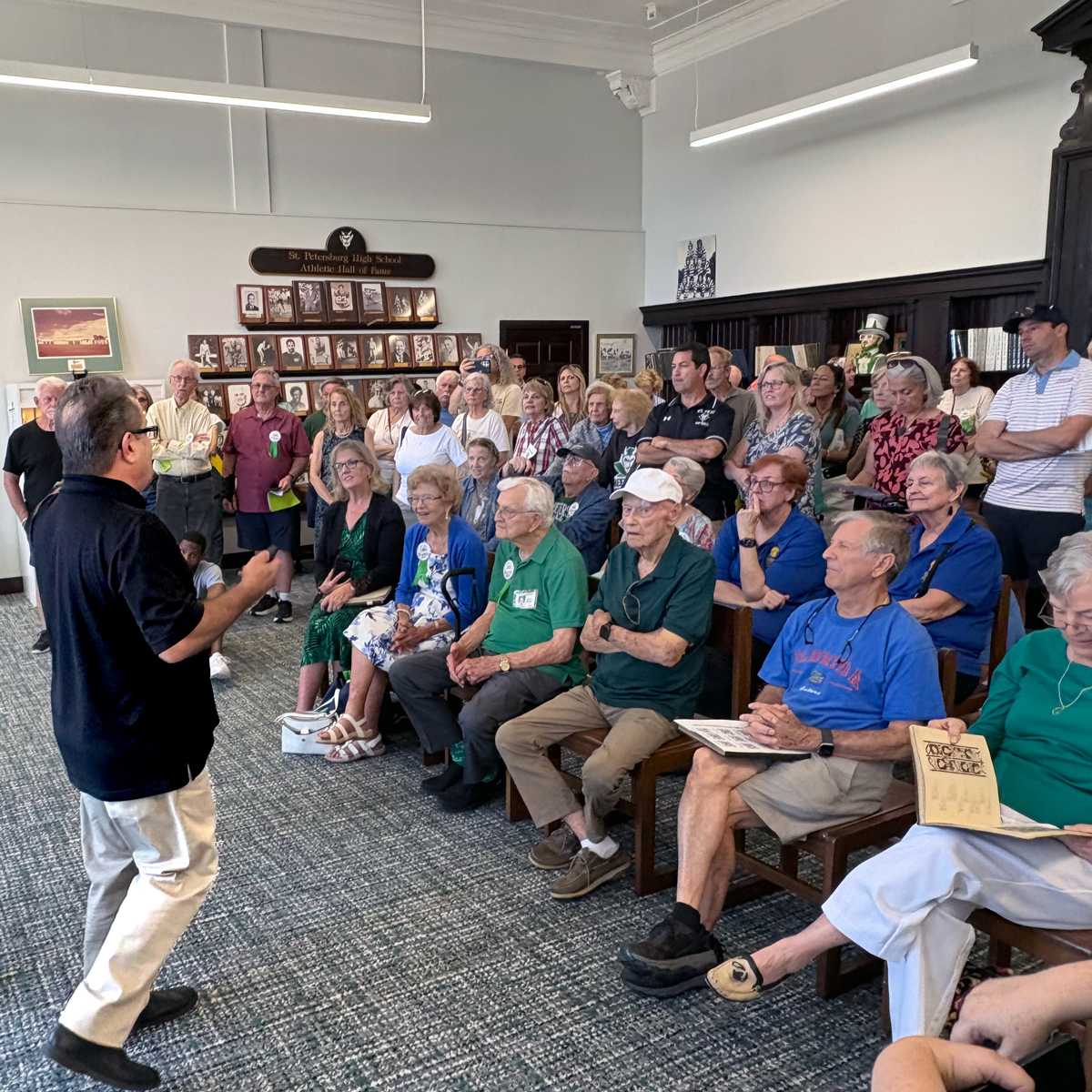 A speaker addresses a large group of seated and standing attendees in the St. Petersburg High School Athletic Hall of Fame room