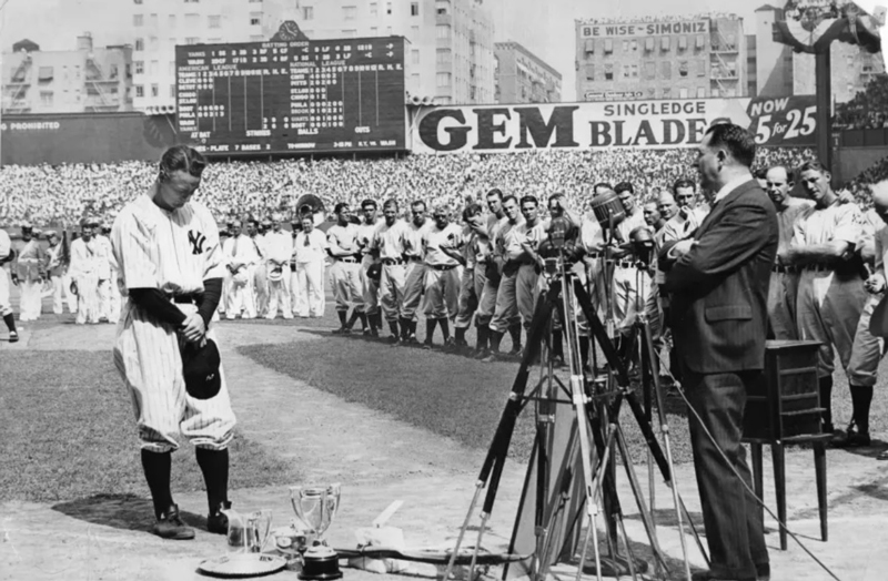 A New York Yankees player bows his head at a microphone ceremony on the field, surrounded by teammates and cameras at a packed stadium