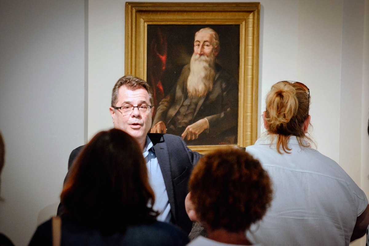 A museum guide speaks to a group of visitors in front of a framed portrait painting of an elderly bearded man
