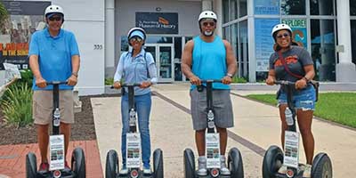 Four people wearing helmets ride Segways in front of the Pensacola Museum of History on a sunny day