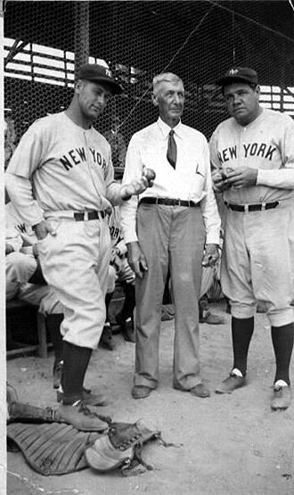 Three men near a baseball dugout, two in New York Yankees uniforms holding bats, posing with a man in street clothes