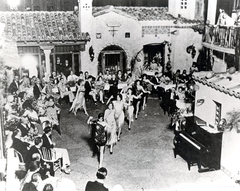 Vintage black-and-white photo of performers dancing in a Spanish-style courtyard venue while guests watch from surrounding tables, with a piano at right