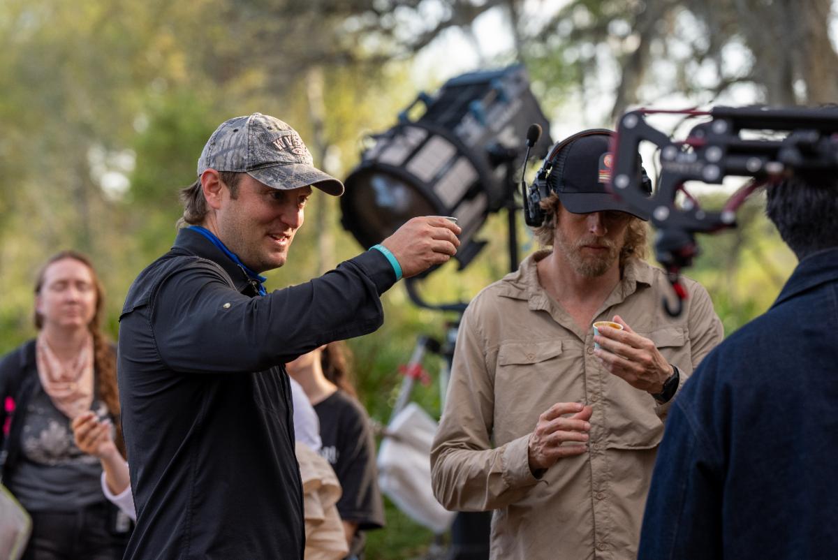 Film crew members on an outdoor set, with lighting equipment and a camera visible in the background