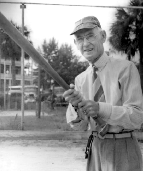 Older man in a cap and tie holding a baseball bat, smiling at the camera, in a black-and-white photo