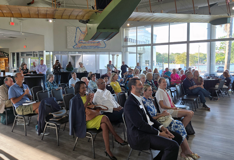A crowd of attendees seated in chairs at an indoor event in a modern, sunlit venue with large windows