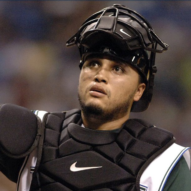 Baseball catcher in full gear, including chest protector and face mask, looking upward during a game
