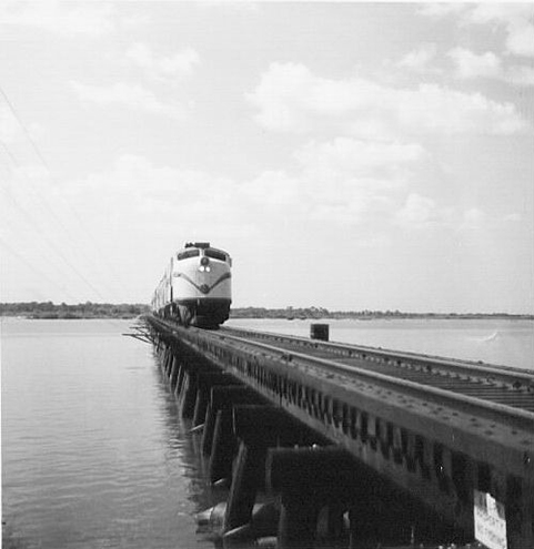 A passenger train crossing a long railroad trestle bridge over calm water in a vintage black-and-white photograph