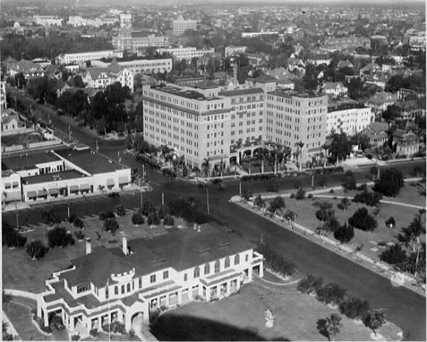 Aerial black-and-white photo of a mid-20th century urban neighborhood featuring a large multi-story hotel and surrounding buildings amid tree-lined streets