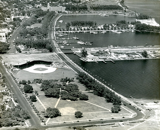 Aerial black-and-white photo of a coastal city with a baseball stadium, marina, and waterfront park