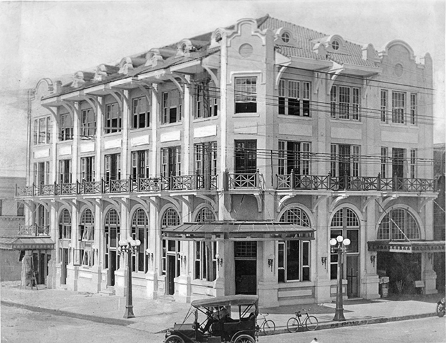 Historic black-and-white photo of a multi-story building with ornate facades, balconies, and arched windows, with an early automobile parked outside