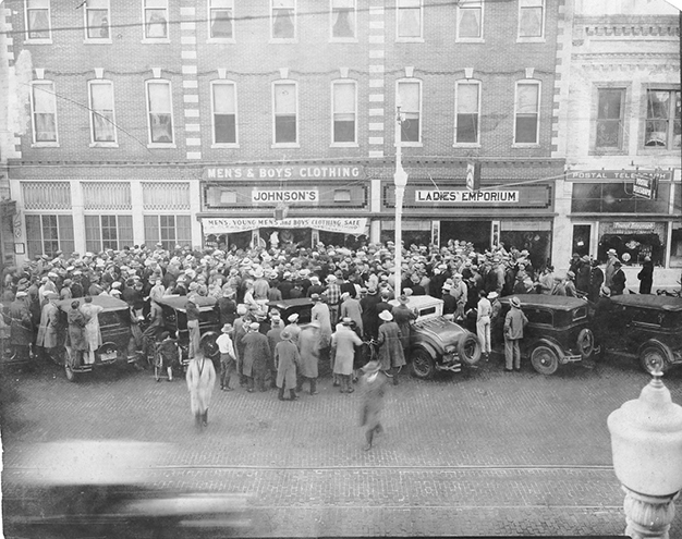 Large crowd gathered outside Johnson's Men & Boys Clothing store and Ladies' Emporium on a city street, circa early 20th century
