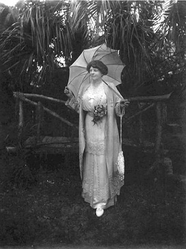 Woman in early 20th-century dress holding a parasol and bouquet, standing outdoors among palm trees