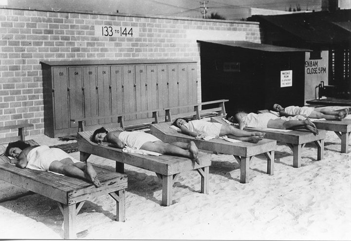 People lying on wooden sunbathing cots in front of numbered lockers 133–144 at a beach facility, vintage black-and-white photo