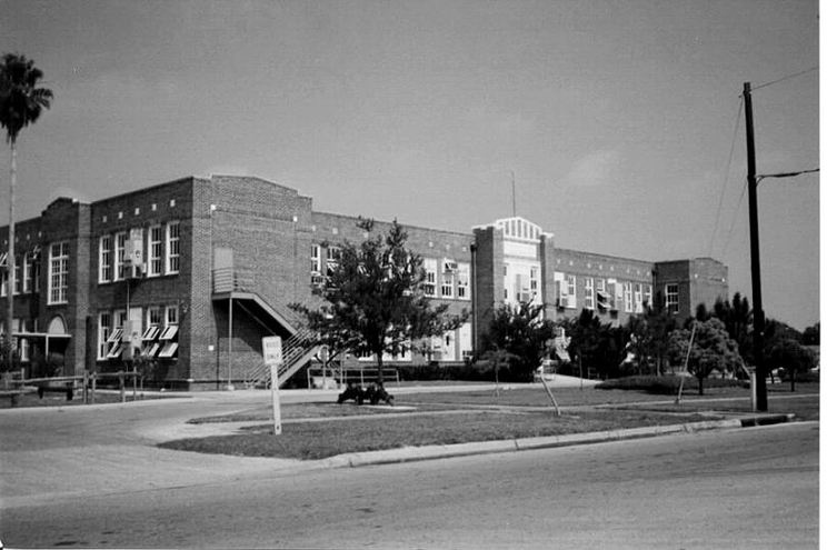 Black-and-white photo of a large brick school building with exterior staircases, trees, and a landscaped traffic circle in the foreground