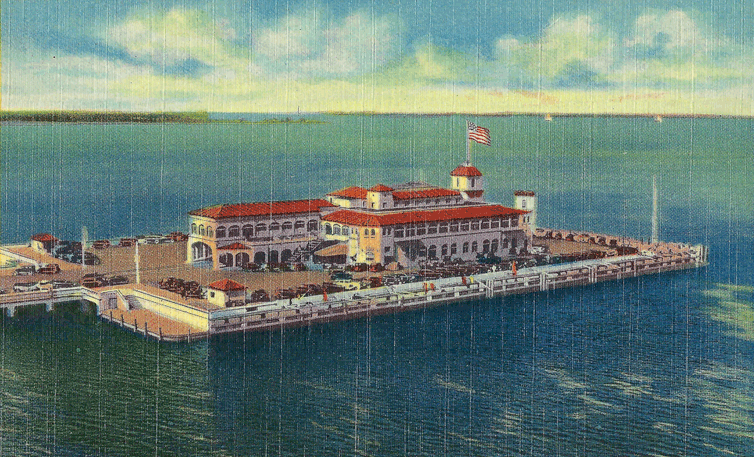 Vintage linen postcard aerial view of a pier with a large Spanish-style building flying an American flag, surrounded by water