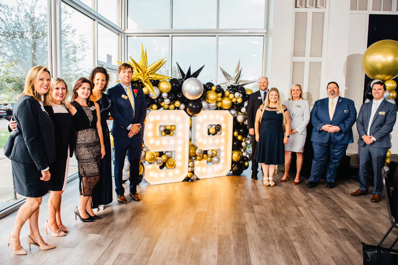 Group of people posing at a 99th anniversary celebration with a lit marquee number 99 and gold, black, and silver balloon decorations