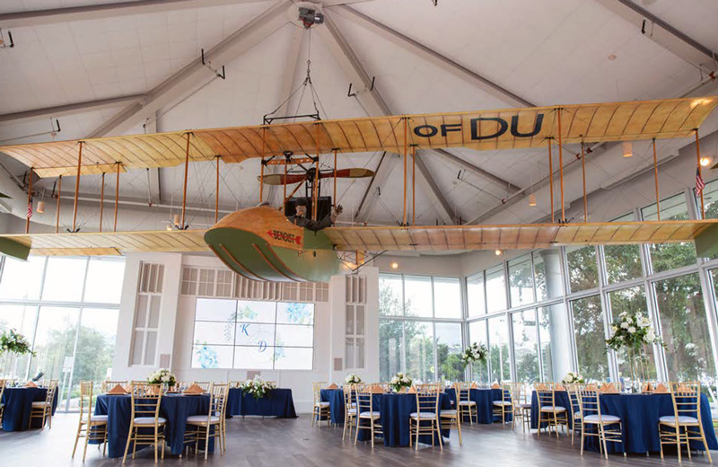 Vintage Benoist biplane suspended from the ceiling above a wedding reception with navy-draped tables and gold chairs