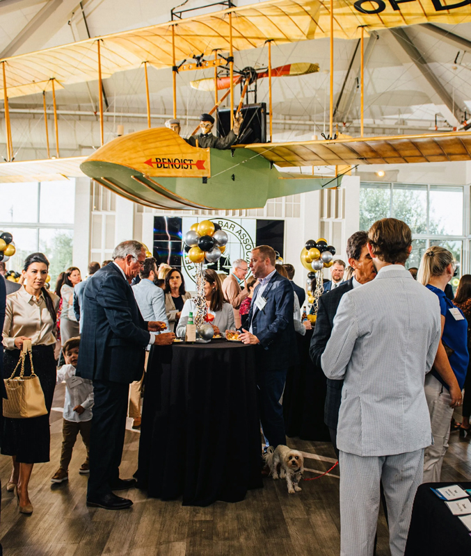Attendees mingle at a Bar Association reception beneath a suspended Benoist aircraft replica in a museum venue