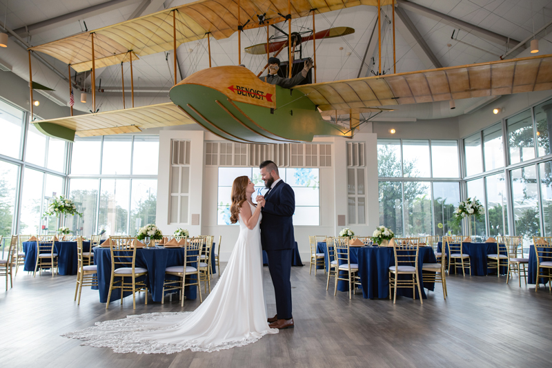 Bride and groom share a first dance beneath a vintage Benoist biplane display in a bright wedding reception venue with navy-draped tables