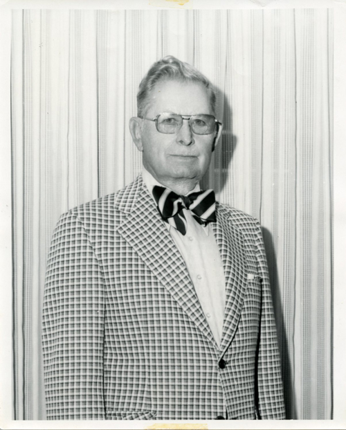 Black-and-white portrait of an older man wearing glasses, a checkered blazer, and a striped bow tie