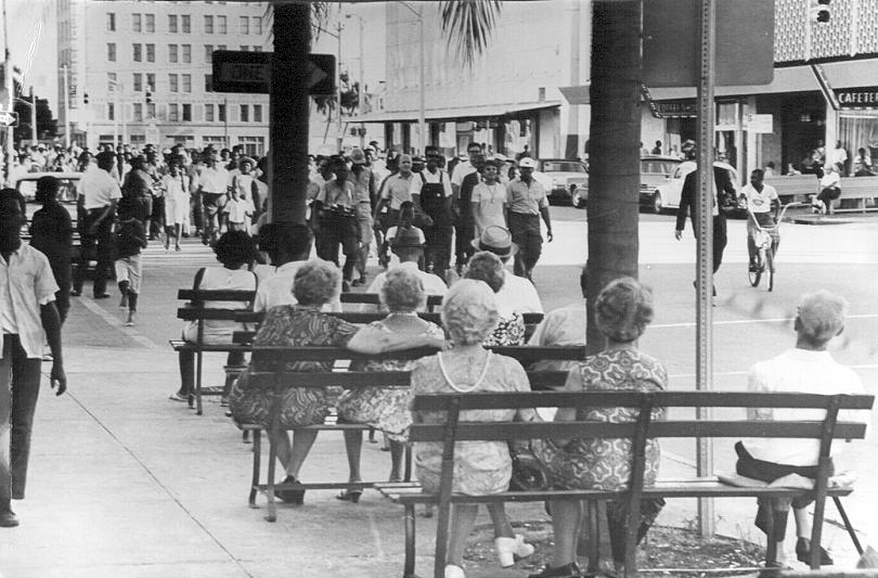 Black and white photo of people seated on sidewalk benches watching a group marching down a city street, likely a civil rights demonstration