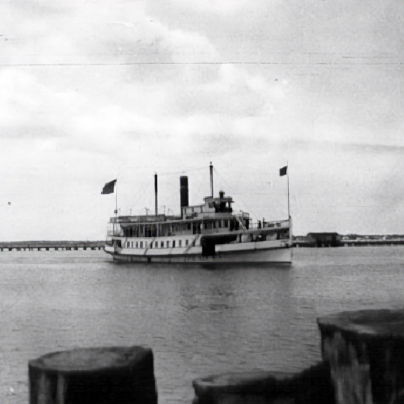 Black-and-white photo of a double-deck paddlewheel steamboat with two smokestacks and flags, on calm water near a pier