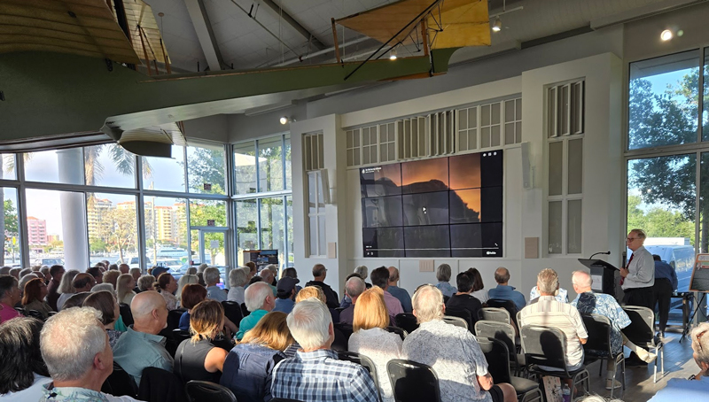 A large audience seated in a bright venue watches a presentation on a large screen, with a speaker at a podium to the right