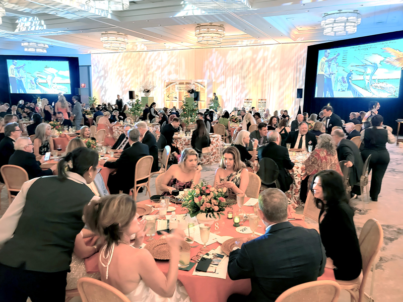 Elegantly dressed guests seated at round tables during a formal gala dinner in a large ballroom with projection screens