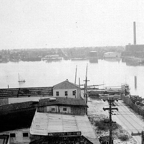 Historic waterfront view with Crescent Tile Co. building, docked boats, and industrial smokestacks across the river