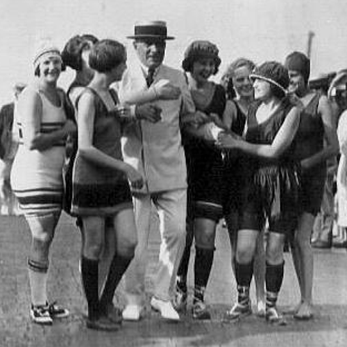A man in a white suit and hat surrounded by women in 1920s-style swimsuits and swim caps outdoors
