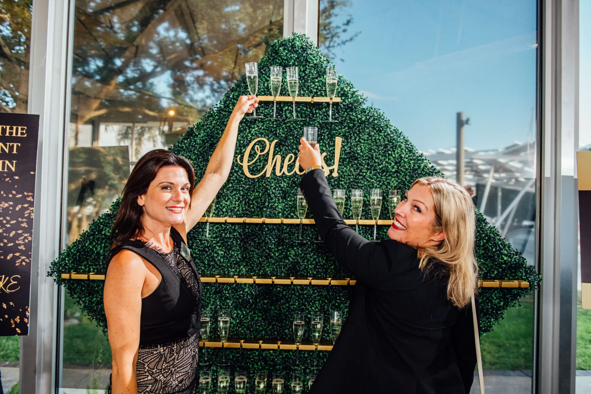Two women toasting with champagne flutes in front of a pyramid-shaped greenery wall display holding champagne glasses, labeled Cheers!