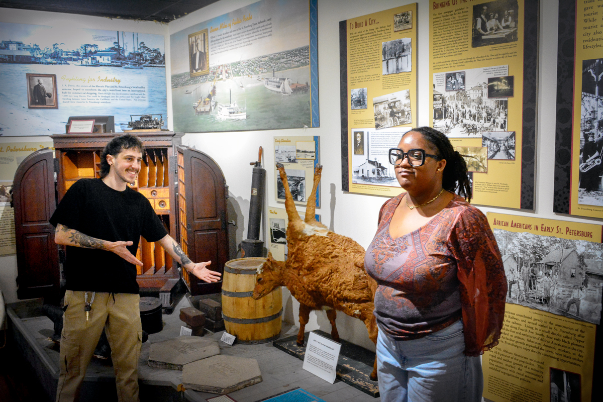 Two visitors exploring a St. Petersburg history museum exhibit featuring a taxidermied animal, antique furniture, and historical panels