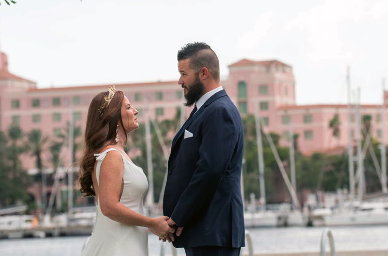 Bride and groom holding hands and facing each other near a marina, with a pink building in the background