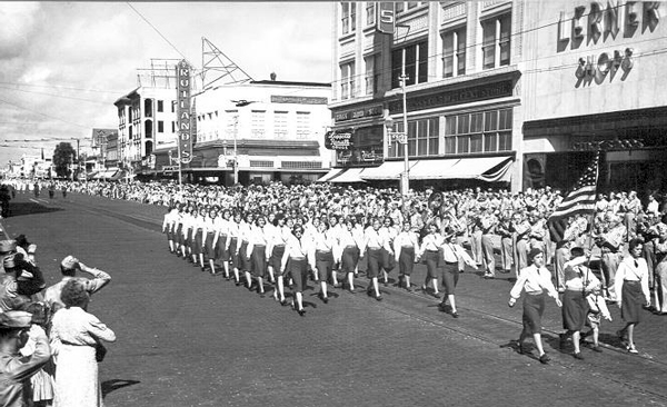 Women in uniform marching in formation down a city street lined with crowds and storefronts, including Lerner Shops, in a mid-20th century parade