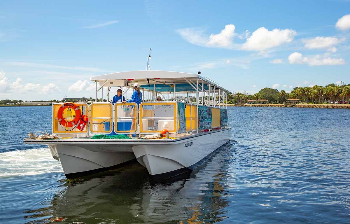 The Discovery boat cruising on calm water with two crew members on deck, palm trees and blue sky in the background