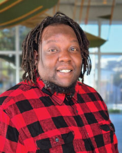 Mario Harris with dreadlocks smiling, wearing a red and black plaid shirt, standing indoors near large windows